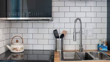A kitchen with a stove, oven, and sink. There is a kettle on the stove, a set of utensils in a holder, and a bottle of dish soap on the counter.