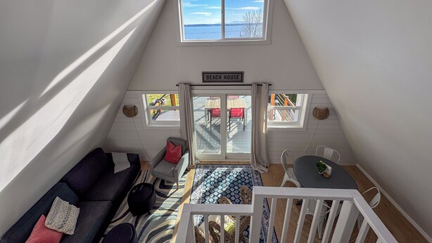 A cozy loft bedroom with a sloped ceiling, a ceiling fan, a large skylight, a view of the outdoors, a bed, a nightstand, and a window with a view of the beach. - Bay View A-Frame + King Bed | 5-Min Walk to Beach (Blaine)