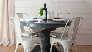 A dining area with a round table, white chairs, a bottle of wine, a small plant, and a woven basket on the wall. - Bay View A-Frame + King Bed | 5-Min Walk to Beach (Blaine)