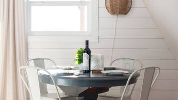 A dining area with a round table, white chairs, a bottle of wine, a small plant, and a woven basket on the wall.