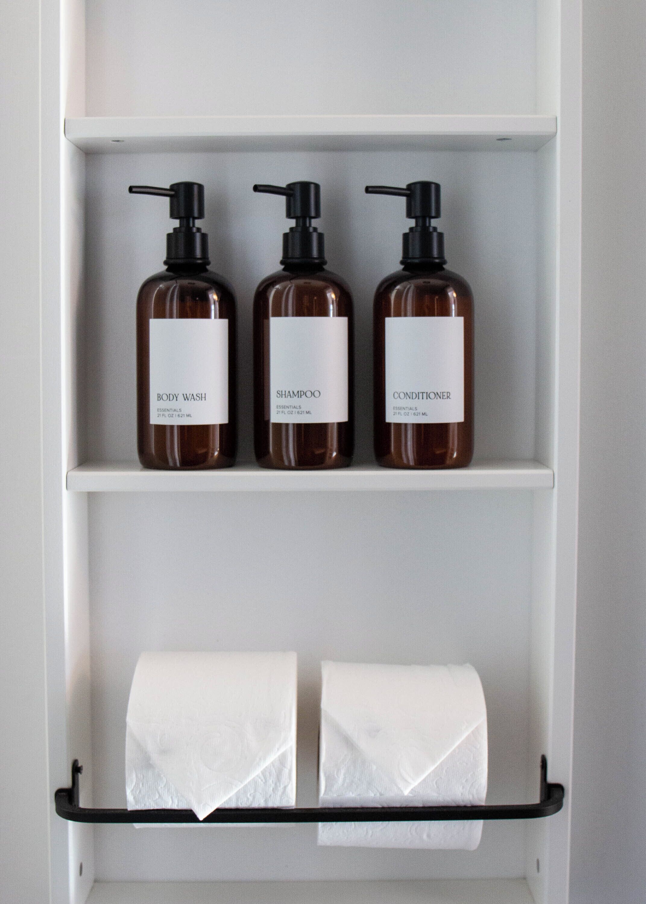 A bathroom shelf with three bottles of body wash, shampoo, and conditioner. Below the shelf, there is a toilet paper holder with two rolls of toilet paper.