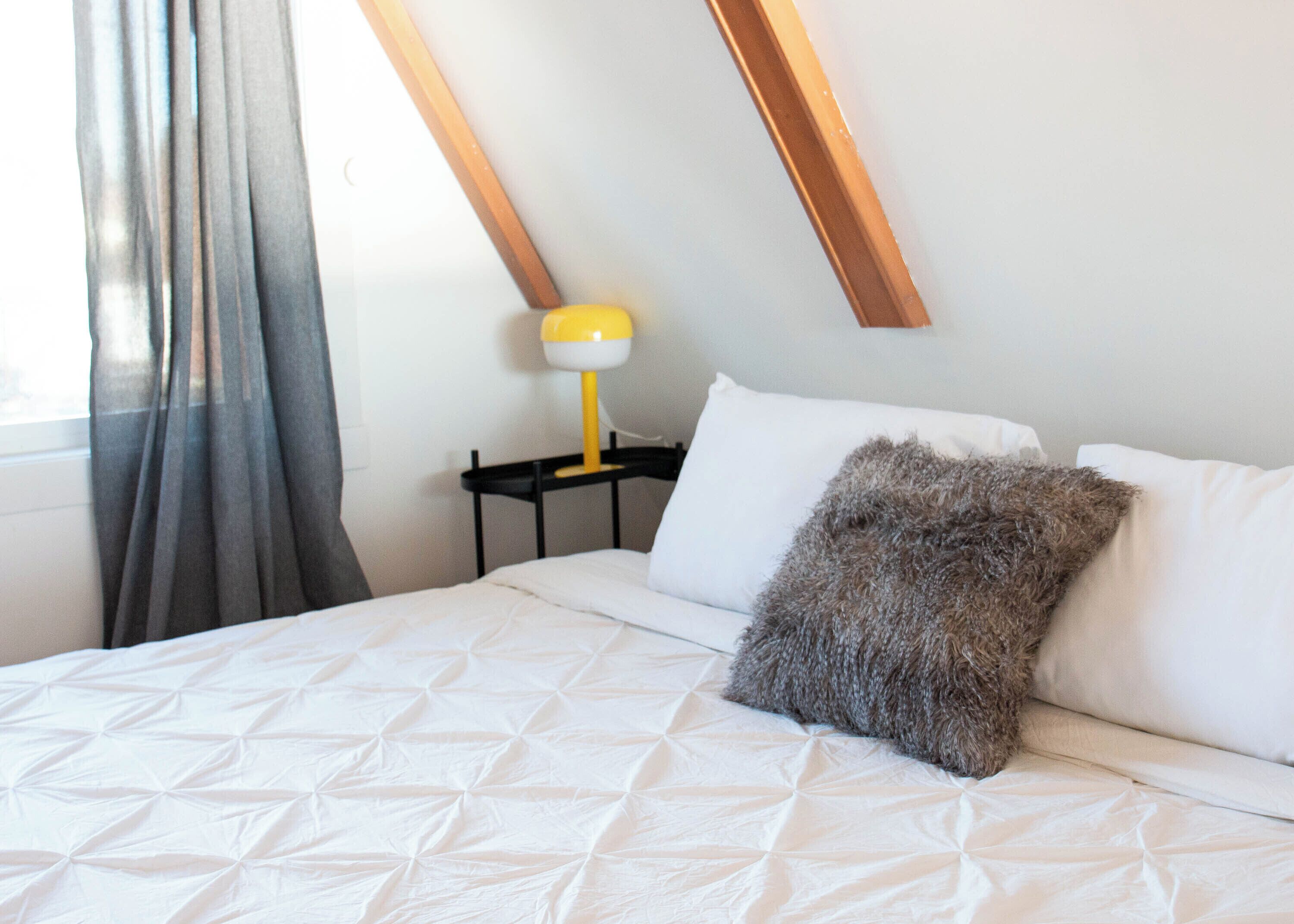 Una cama bien hecha con colcha blanca, un almohadón mullido y una mesita de noche con una lámpara amarilla.