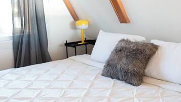 Una cama bien hecha con colcha blanca, un almohadón mullido y una mesita de noche con una lámpara amarilla.