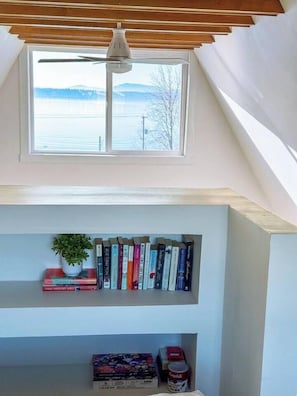 A room with a sloped ceiling, a skylight, a ceiling fan, a bookshelf with books and a plant, and a window with a view of the outdoors. - Bay View A-Frame + King Bed | 5-Min Walk to Beach (Blaine)