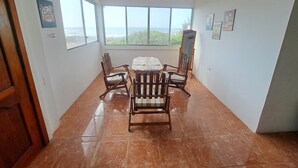 A dining area with a large table and six wooden chairs, large windows with a view of the ocean, and a tiled floor.