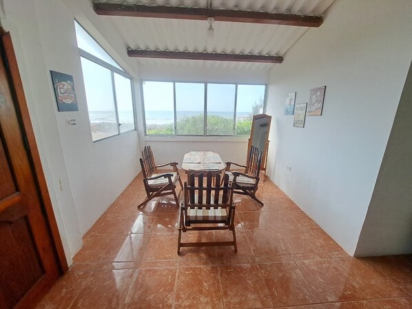 Un comedor con una mesa grande y seis sillas de madera, ventanas amplias con vista al océano y piso de baldosas.