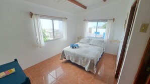 A bedroom with a bed, two windows with curtains, a small table, and a door.