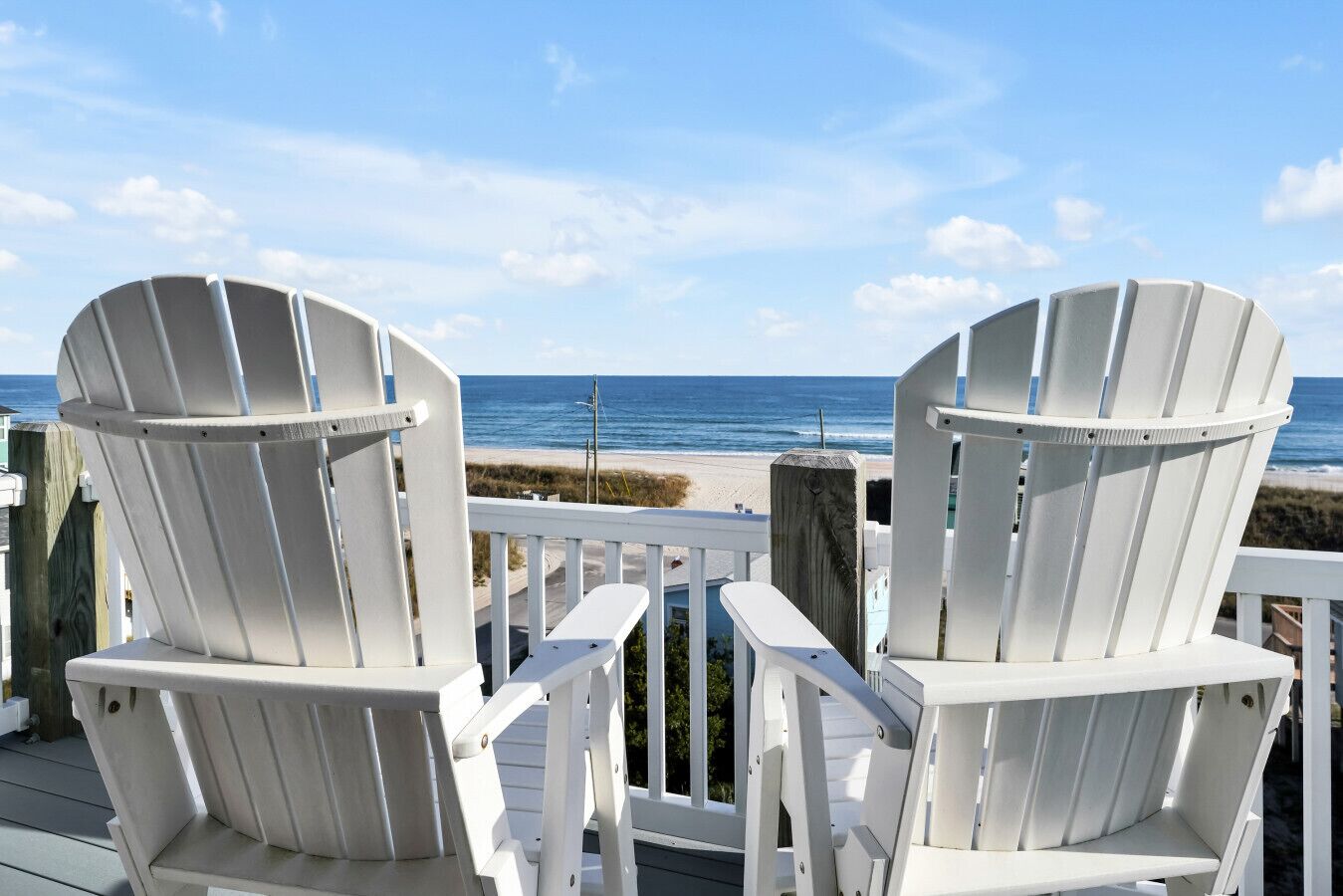 Two white Adirondack chairs facing a beach with a wooden railing in the foreground.