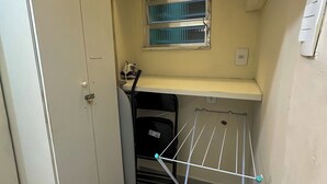 Una habitación pequeña con un armario blanco, un ventanal con persianas, un tendedero y una maleta negra sobre una estantería.