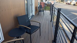 Un balcon avec deux chaises et une table, donnant sur une rue bordée de voitures garées et d’immeubles.