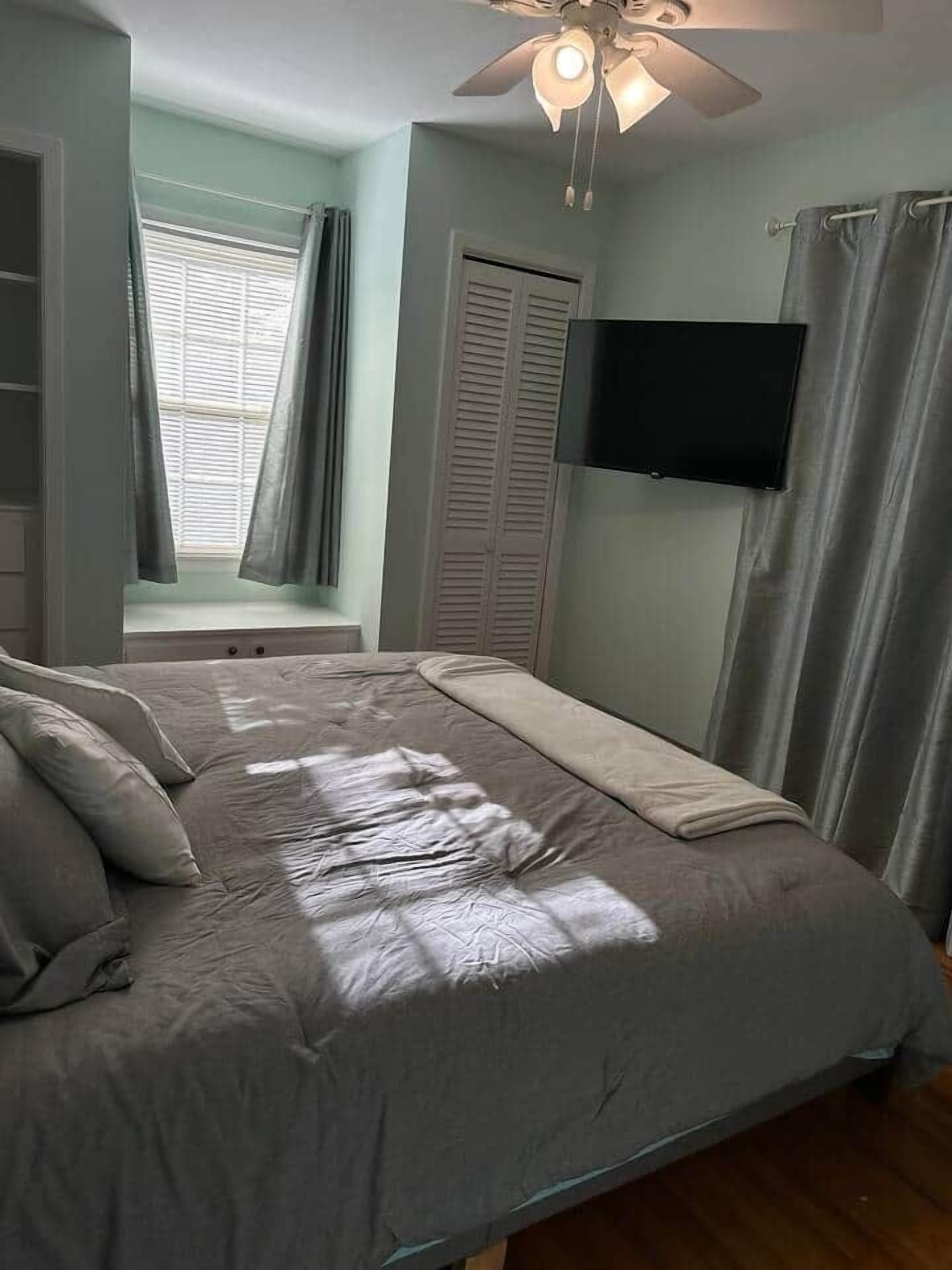Ein Schlafzimmer mit Bett, Deckenventilator, Fenster mit Vorhängen, Wandfernseher und Kleiderschrank.