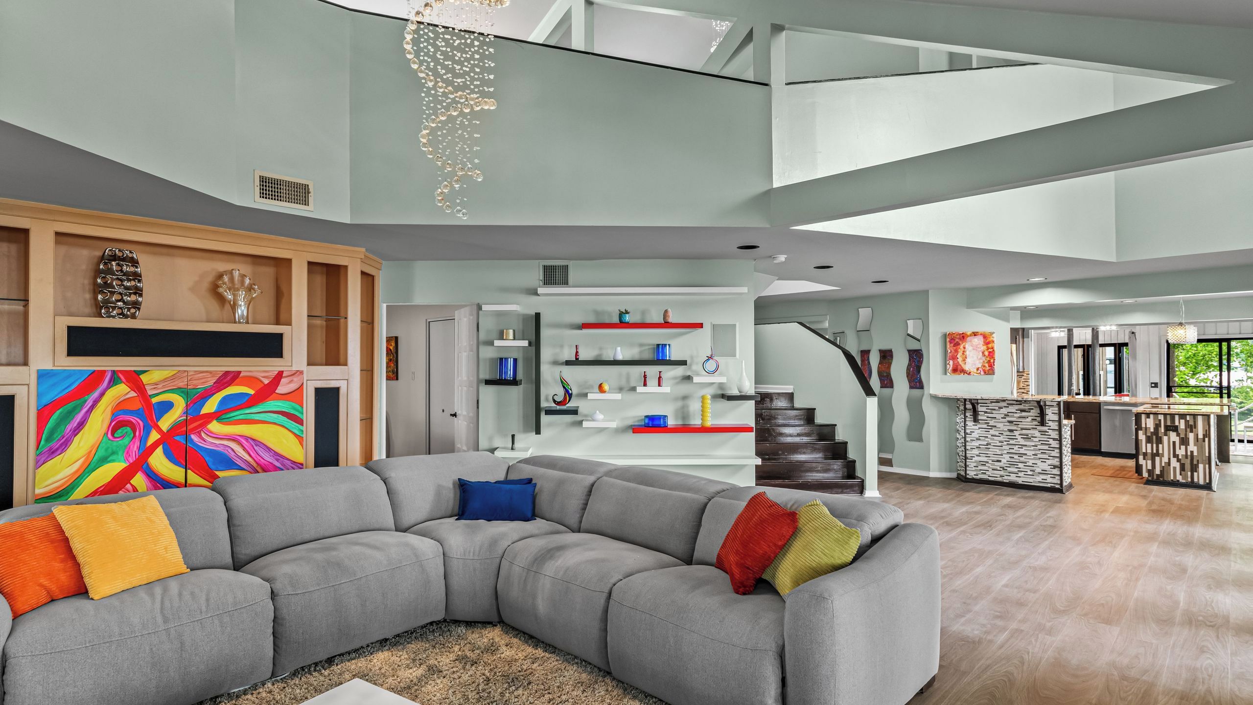 A modern living room with a sectional sofa, a colorful abstract painting, and a staircase leading to an upper level.