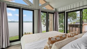 A bedroom with a large window overlooking a lake, a bed with white bedding, and a view of a deck with a bird. - Waterfront Stay w/ Canoes, Deck, Game Room & Lake Views Near Mooresville Renwick Cove by Avantstay (Mooresville)