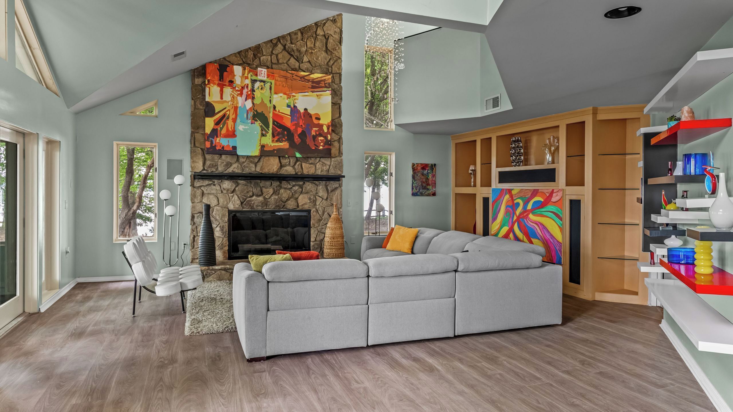 A modern living room with a stone fireplace, a grey sectional sofa, and colorful abstract art.
