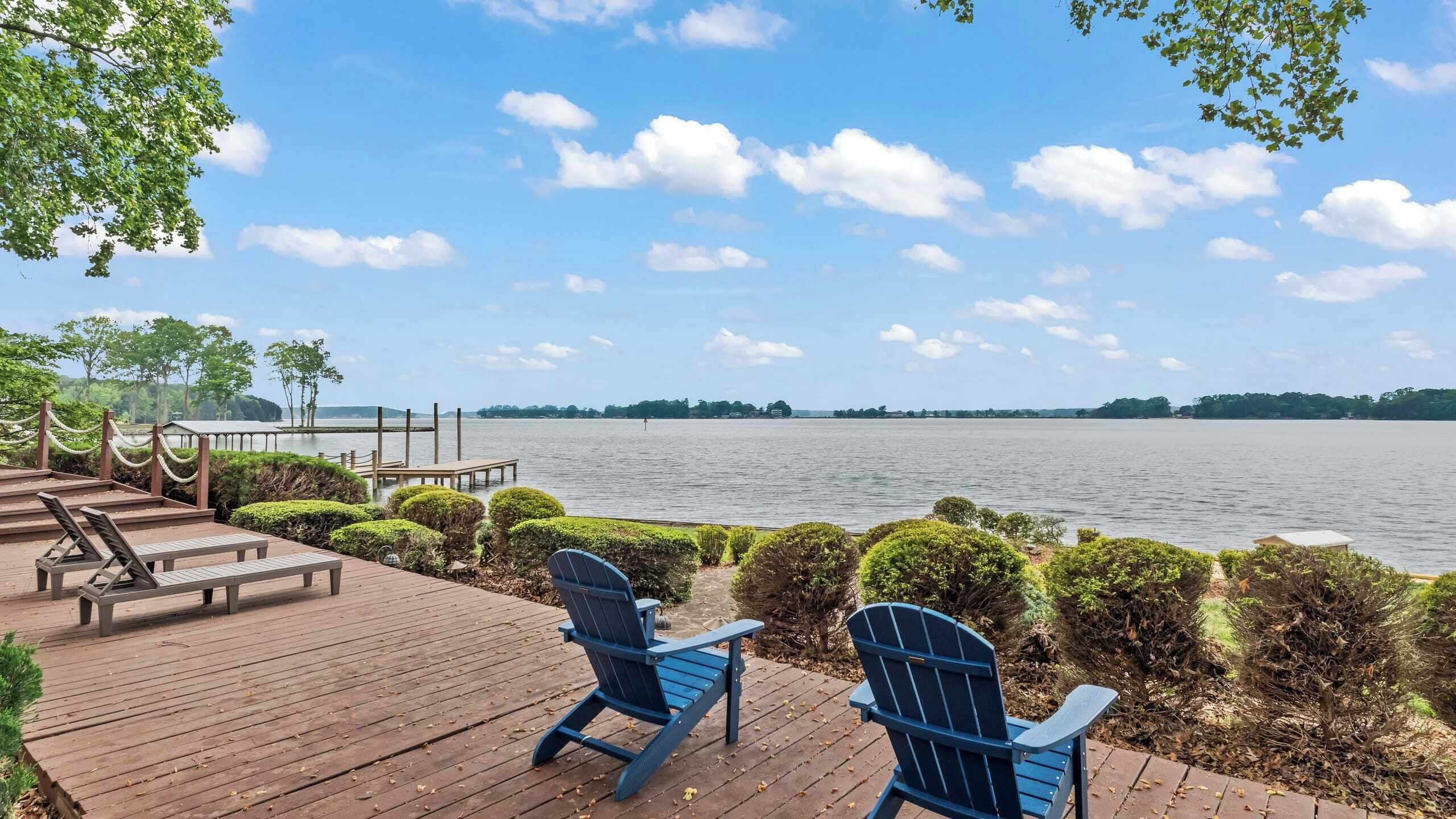 Une terrasse en bois avec deux fauteuils Adirondack donnant sur un lac, entourée de buissons soigneusement taillés et d’une barrière faite de cordes.