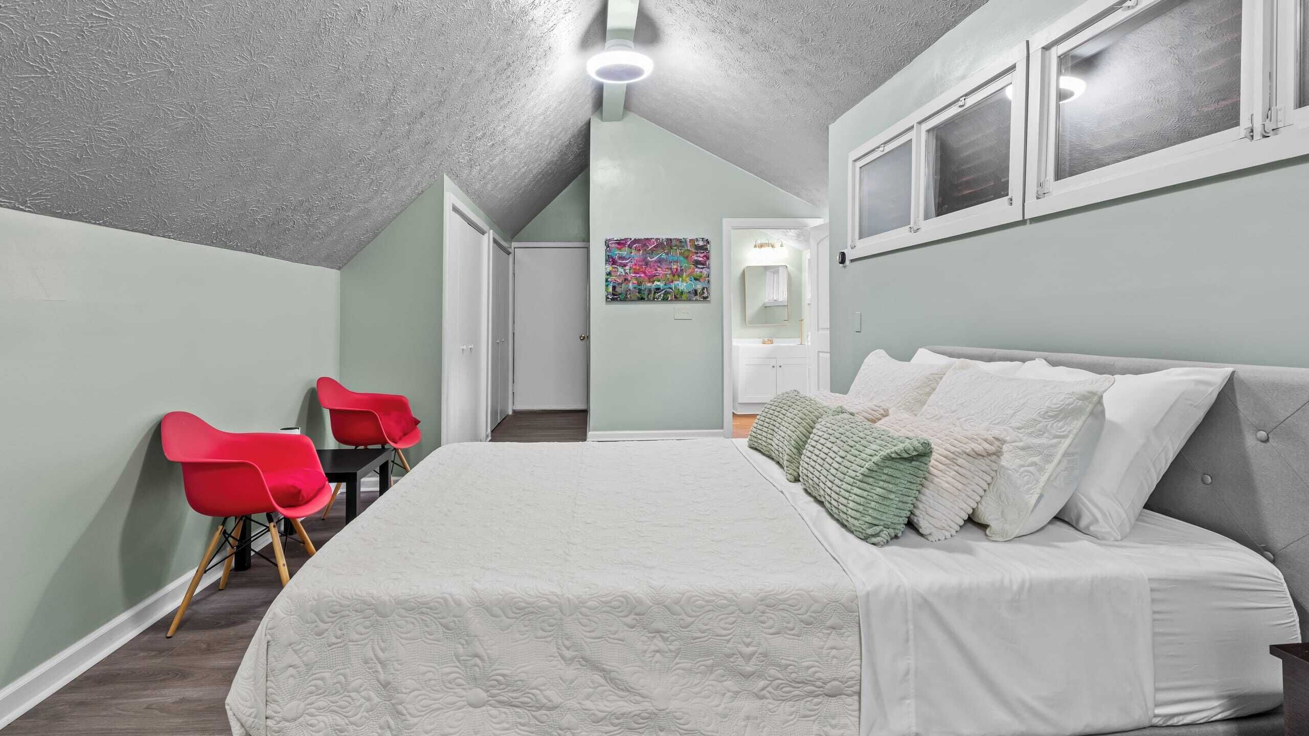 Ein Schlafzimmer mit einem großen Bett, einem Schreibtisch mit zwei roten Stühlen, einem Gemälde an der Wand und einem Fenster mit Jalousien.