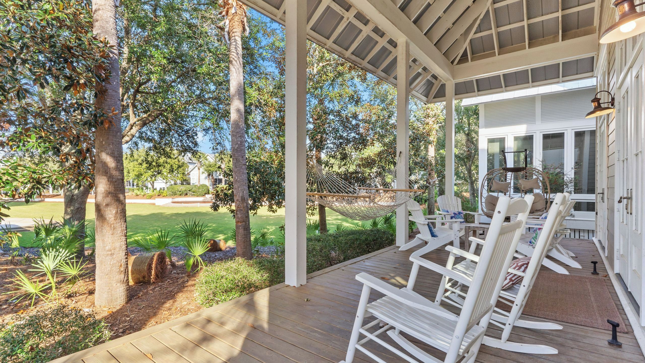 A covered outdoor seating area with white chairs and a table, surrounded by trees and a lawn.