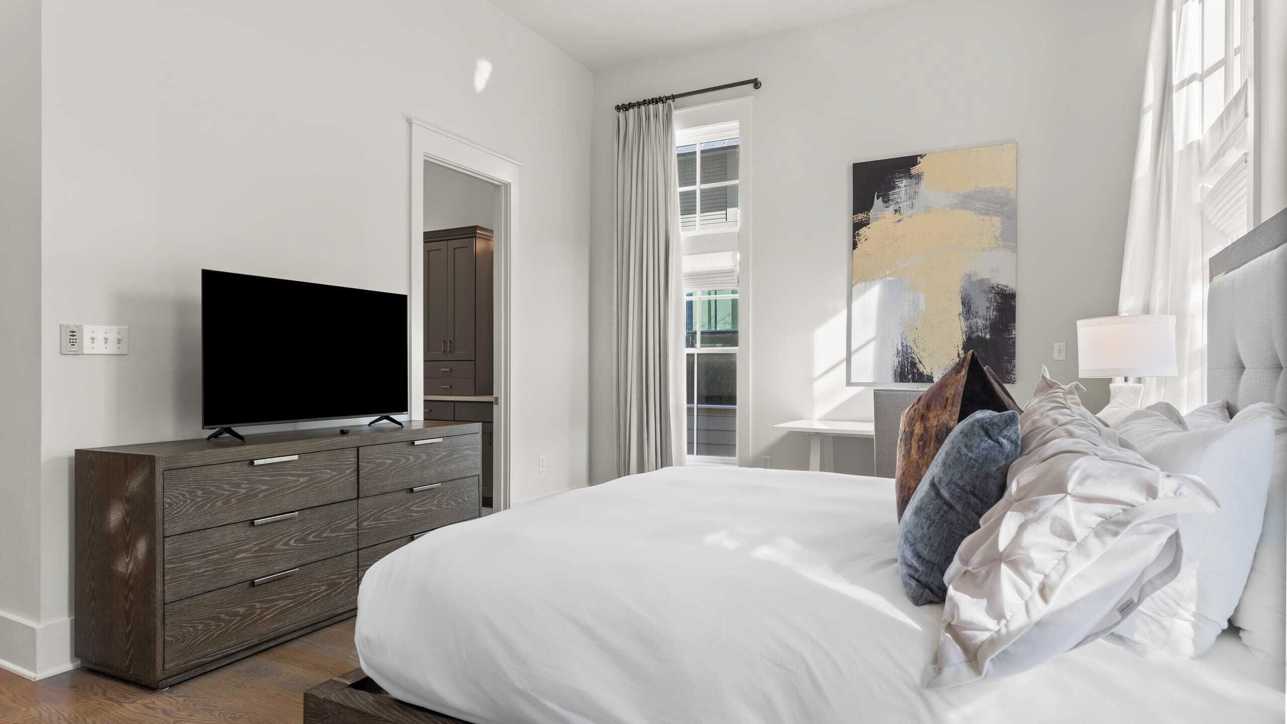 A modern bedroom with a large bed, a TV on a wooden dresser, and a painting on the wall.