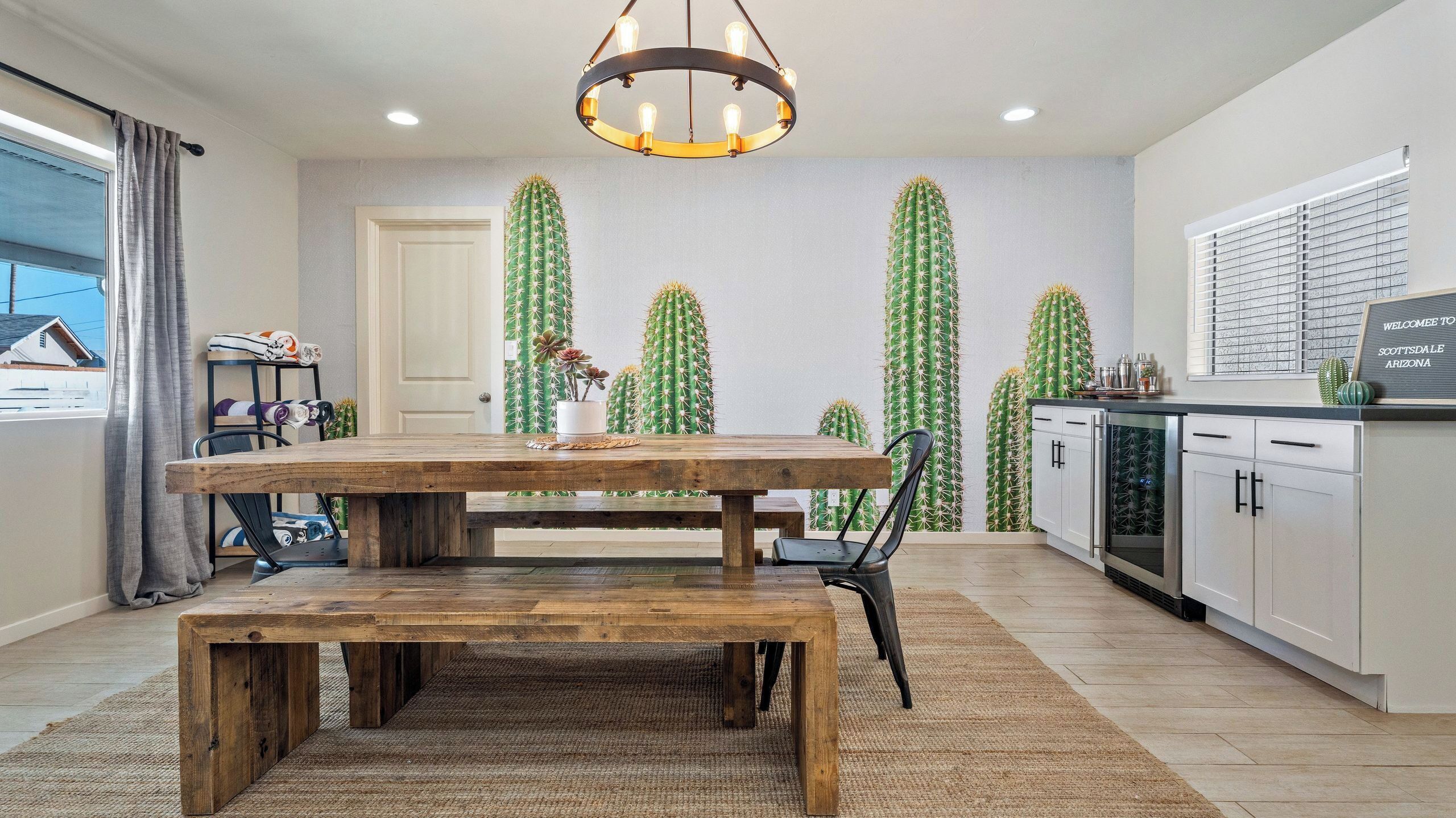 Un coin repas avec une table et des bancs en bois, un lustre, et une cuisine dotée de meubles blancs et d’un revêtement mural.