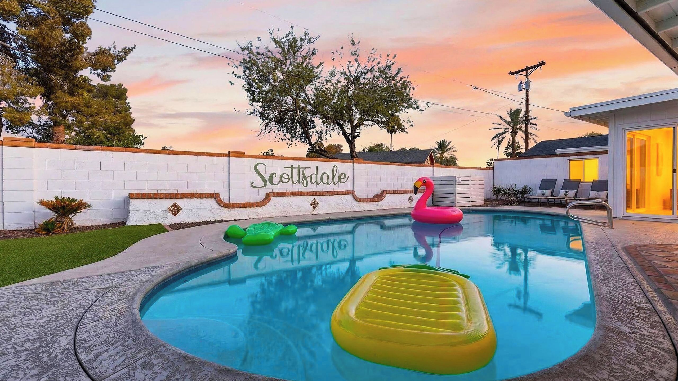 A swimming pool with inflatable toys, a white wall with 'Scottsdale' written on it, and a sunset in the background.