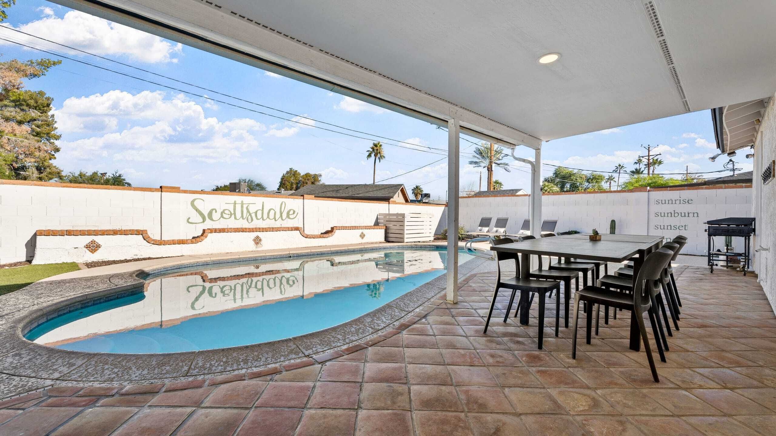 An outdoor pool area with a covered patio, a table with chairs, and a wall with the text "Scottsdale Sunrise Sunset".