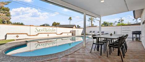 An outdoor pool area with a covered patio, a table with chairs, and a wall with the text "Scottsdale Sunrise Sunset".
