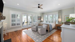 A bedroom with a bed, two chairs, a desk, a ceiling fan, and large windows. - Beachside Stay With Pool, Two Decks, Rooftop Lounge & Steps to Front Beach IOP Beachhouse by Avantstay (Isle of Palms)