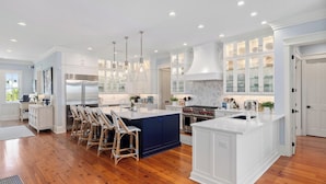 A spacious kitchen with white cabinetry, a large island, and a dining area with wicker chairs. - Beachside Stay With Pool, Two Decks, Rooftop Lounge & Steps to Front Beach IOP Beachhouse by Avantstay (Isle of Palms)