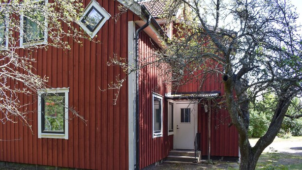 Un bâtiment en bois rouge, doté d’une porte et de fenêtres, entouré d’arbres et d’herbe.
