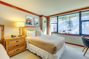 Habitación de hotel con cama, mesita de noche con lámpara, silla y ventana con vistas a un paisaje nevado.