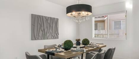 Ein Esszimmer mit Holztisch, grau gepolsterten Stühlen, einer Chandelüstere, einem Wandbild im abstrakten Stil und einem Fenster mit Blick auf ein Gebäude.