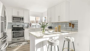A modern kitchen with white cabinetry, stainless steel appliances, a central island with stools, and a view of the outdoors. - Vista del Sol by Avantstay Bright Hilltop Stay + Patio + Views + Premium Kitchen & Space for Groups (San Diego)