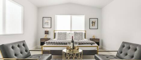 Quarto moderno com uma cama grande, duas mesas de cabeceira, uma mesinha de apoio dourada e duas cadeiras estofadas cinzas.