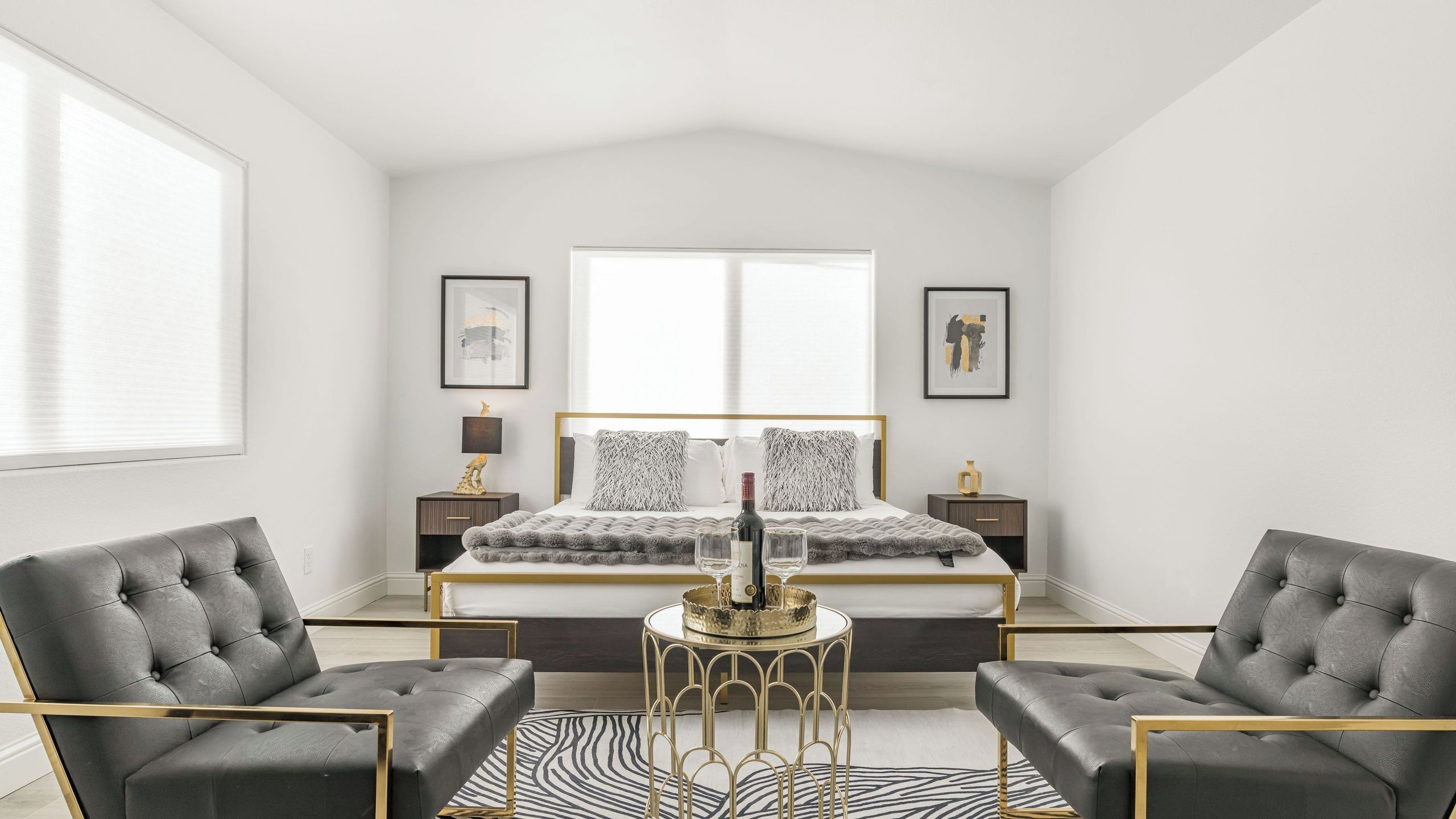 A bedroom with a bed, two nightstands, a table with a bottle and glasses, two armchairs, and a window.
