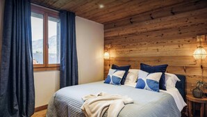 A bedroom with a wooden ceiling, a large bed with blue and white bedding, a window with curtains, and two wall-mounted lamps. - Chalet Trumont - Modern comfort and Alpine charm - OVO Network (Samoëns)
