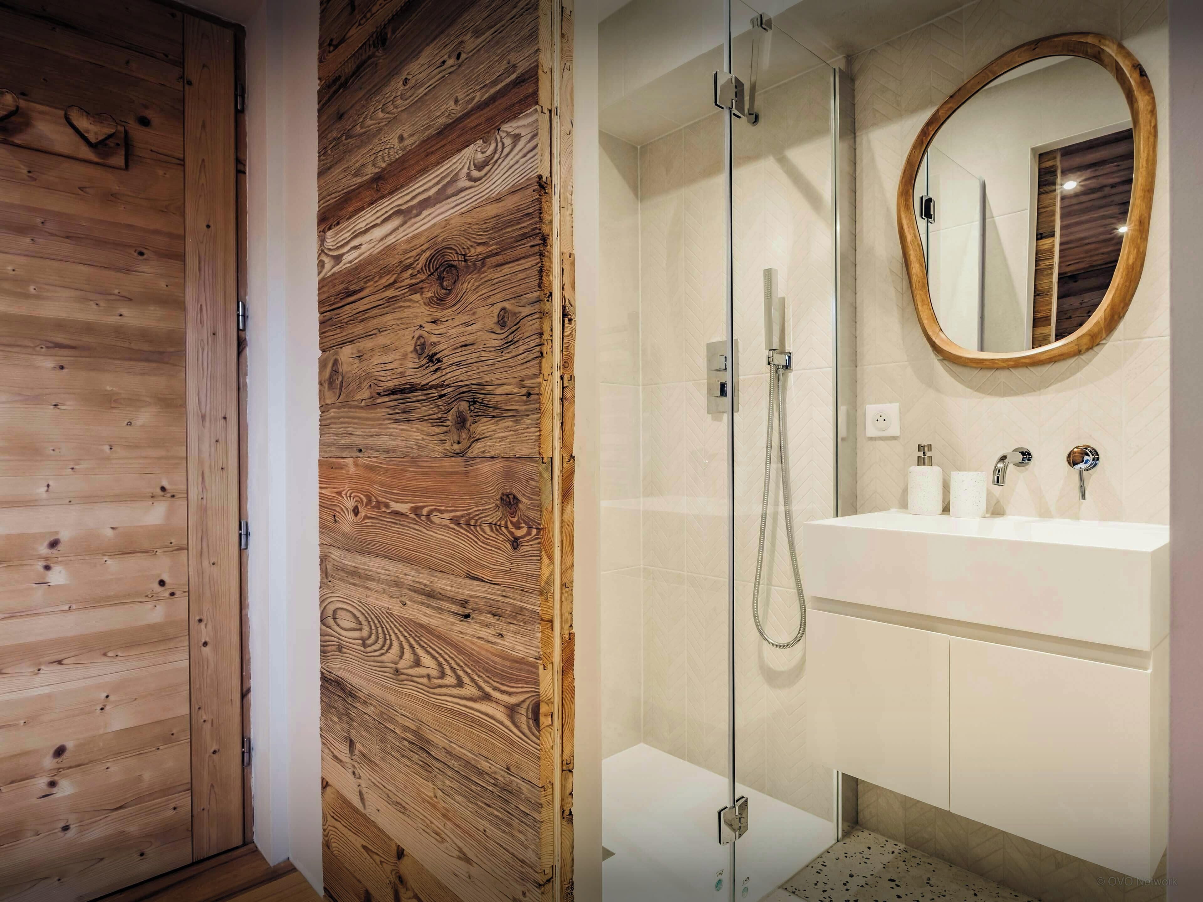 Ein Badezimmer mit Holztür, einer Duschkabine aus Glas, einem weißen Waschbecken und einem Spiegel mit Holzrahmen.