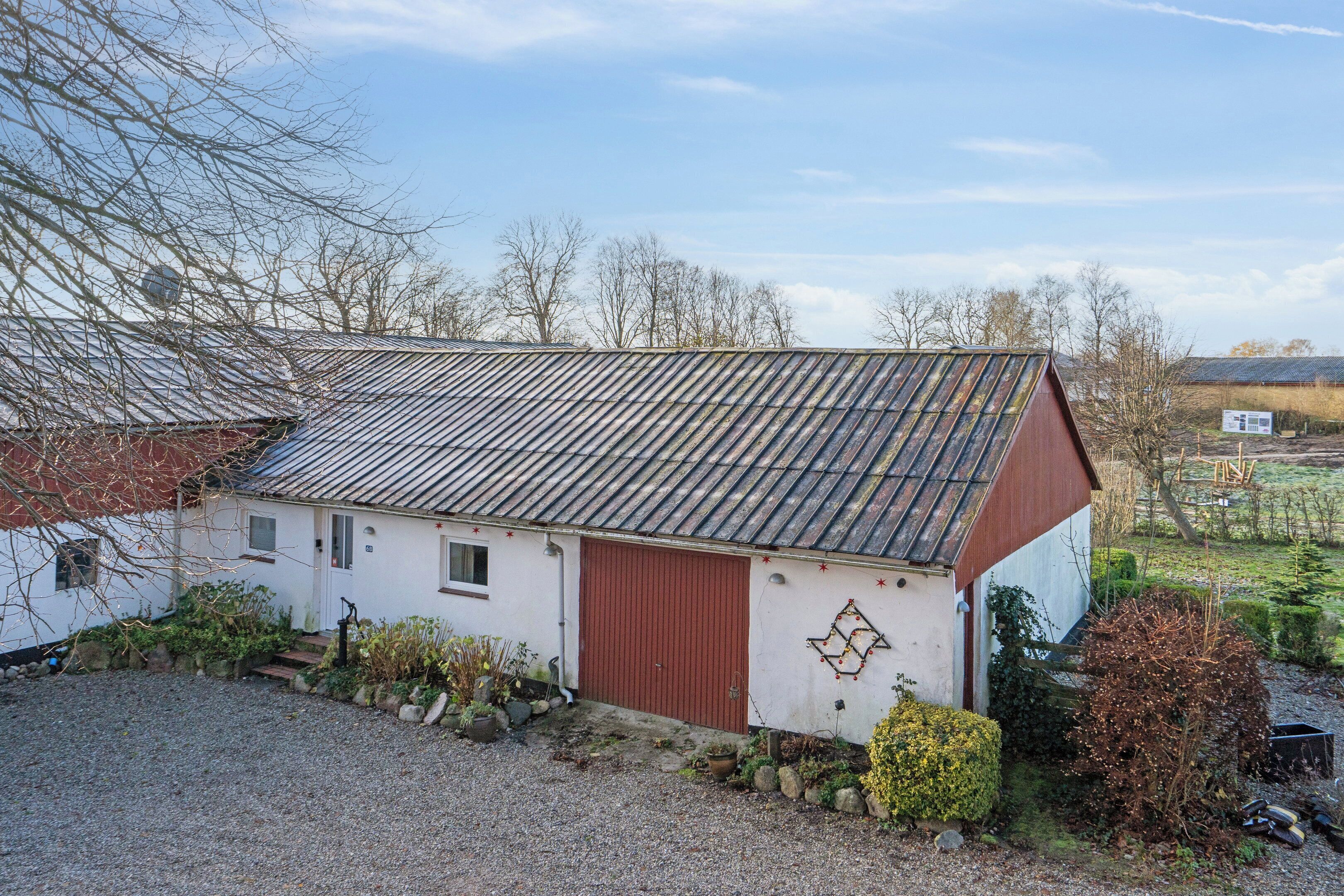 A small, single-story building with a red door and a sloped metal roof, surrounded by a gravel driveway and sparse vegetation.