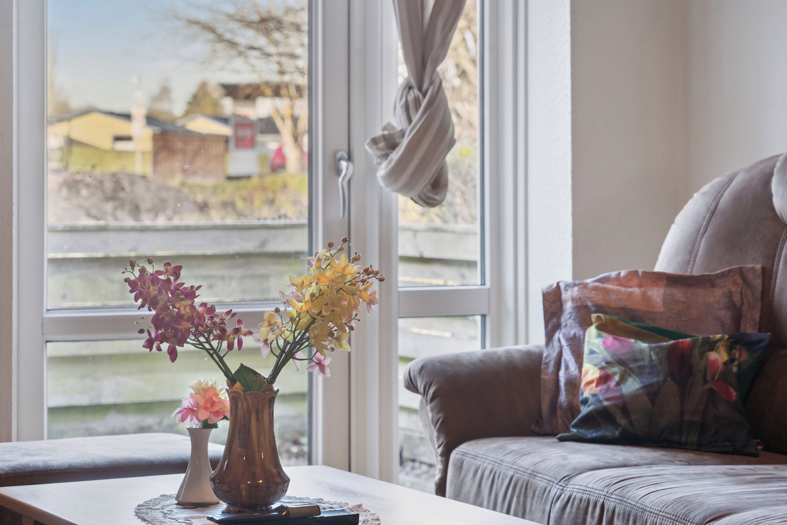 A room with a window, a table with a vase of flowers, and a sofa with a colorful pillow.