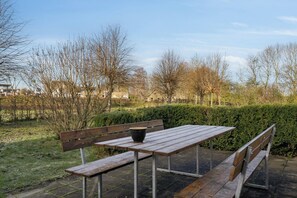 An outdoor seating area with a wooden bench and table, surrounded by trees and a hedge.