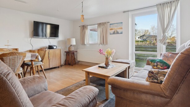 A living room with a sofa, a coffee table, a TV, a dining area with a table and chairs, and a view through a large window.