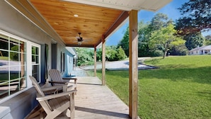 Une terrasse en bois couverte, dotée de chaises Adirondack, d’un ventilateur de plafond et offrant une vue sur une pelouse et des arbres.