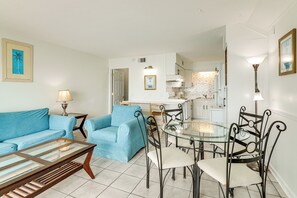 A living room with a blue sofa, a glass coffee table, and a dining area with a glass table and metal chairs. - Steps to Pools & Beach! 11th-Floor PCB Retreat (Panama City Beach)