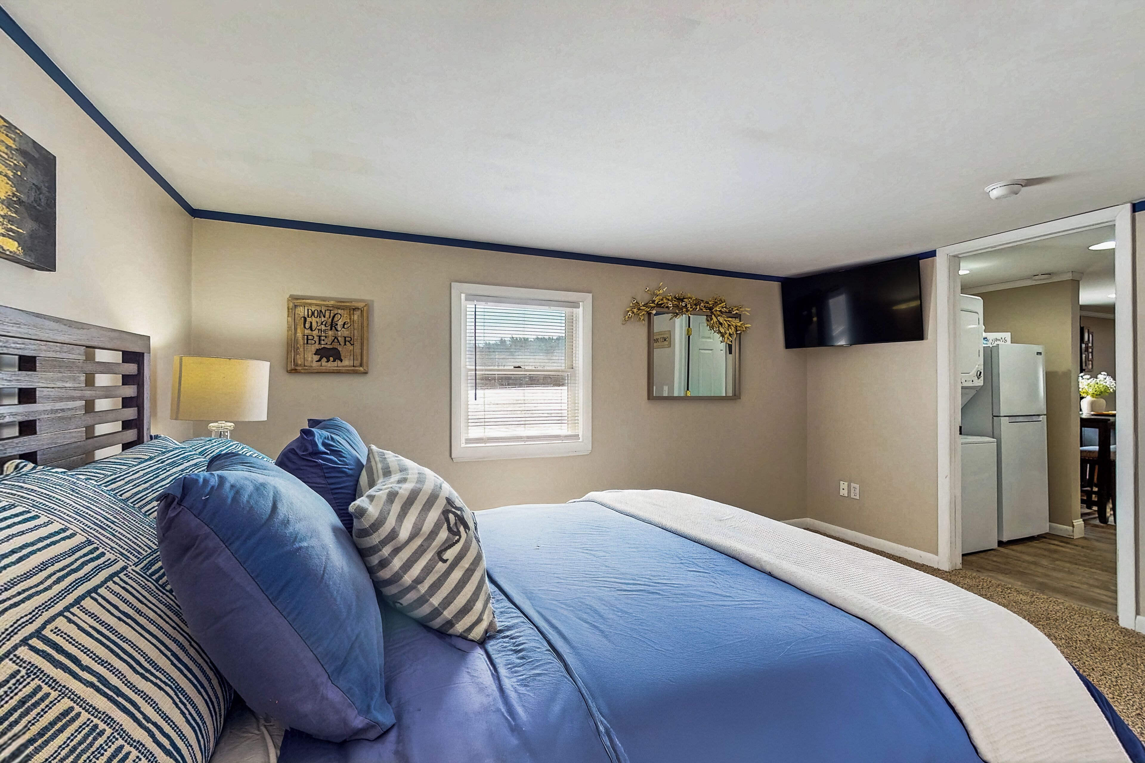 A bedroom with a bed, pillows, a bedside table, a window, and a door leading to a kitchen.