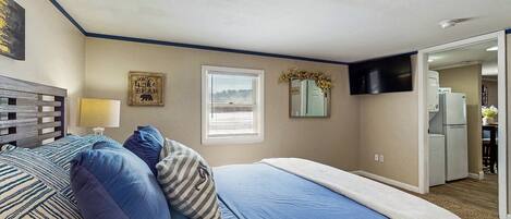 A bedroom with a bed, pillows, a bedside table, a window, and a door leading to a kitchen.
