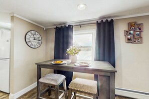 A small kitchen with a wooden table, two stools, a clock, and a window with curtains.