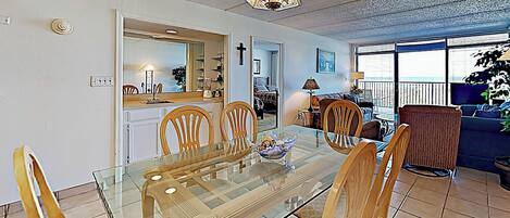 A dining area with a glass table and wooden chairs, a kitchen in the background, and a balcony with a view.