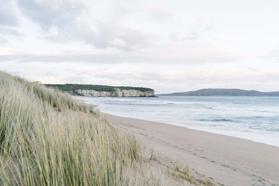 A beach with a grassy dune, a sandy shore, and a cliff in the background.