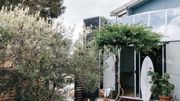 Una casa moderna con fachada de vidrio, terraza de madera y una tabla de surf apoyada en la puerta.