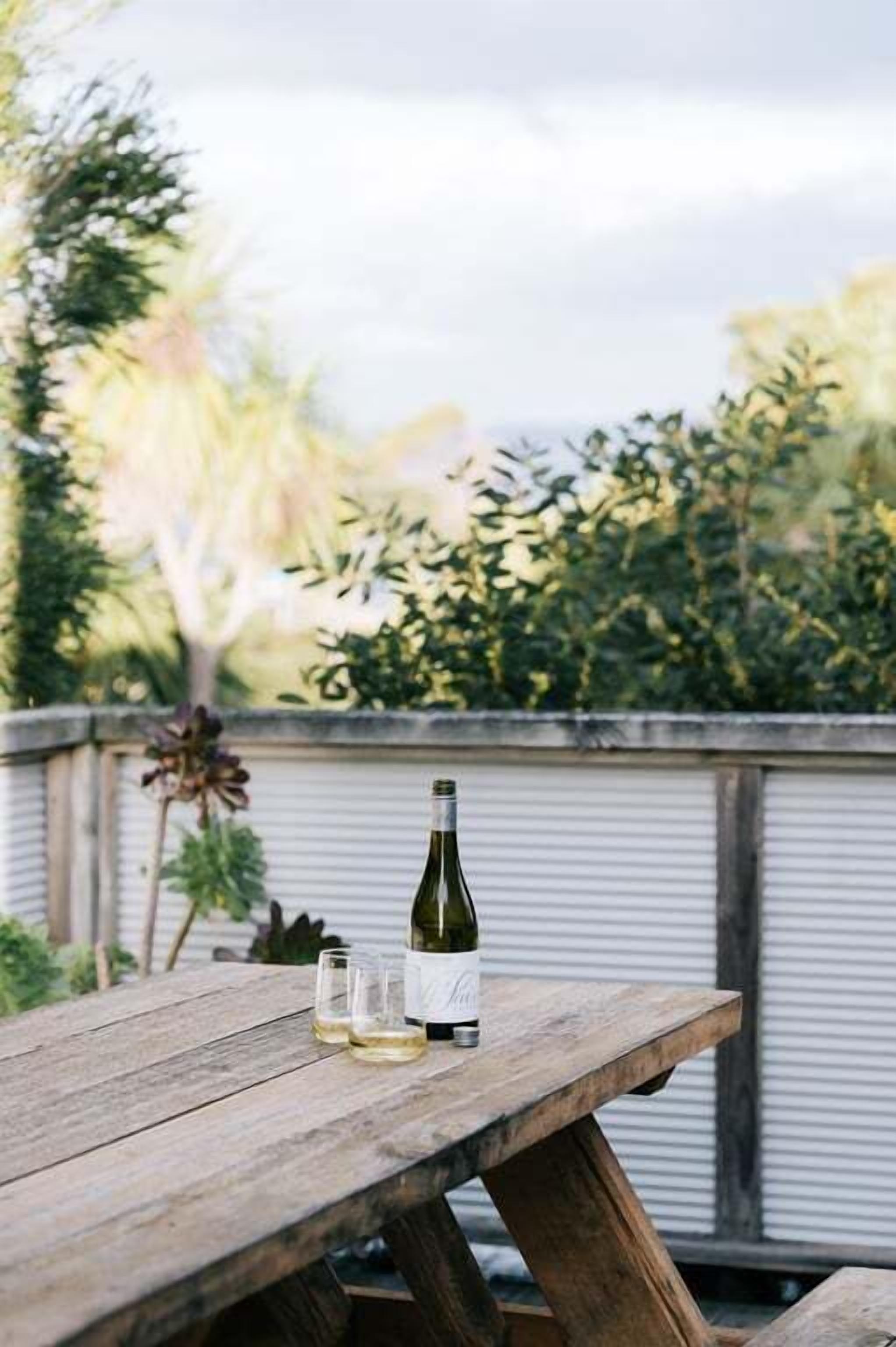A wooden picnic table with a wine bottle and two glasses on it, set outdoors with a view of greenery and a metal fence in the background.