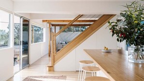 Una cocina moderna con una escalera de madera, una zona de comedor con taburetes y un amplio ventanal que proporciona luz natural.